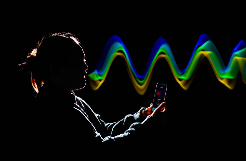 A silhouetted person holds a smartphone covered in rim lighting, appearing to record or speak into it while luminous green, yellow, and blue waves emanate in front of them, suggesting sound or data transmission.